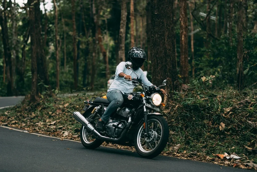Homem pilotando uma moto Royal Enfield preta em estrada asfaltada no meio da floresta tropical, usando capacete e camisa clara.