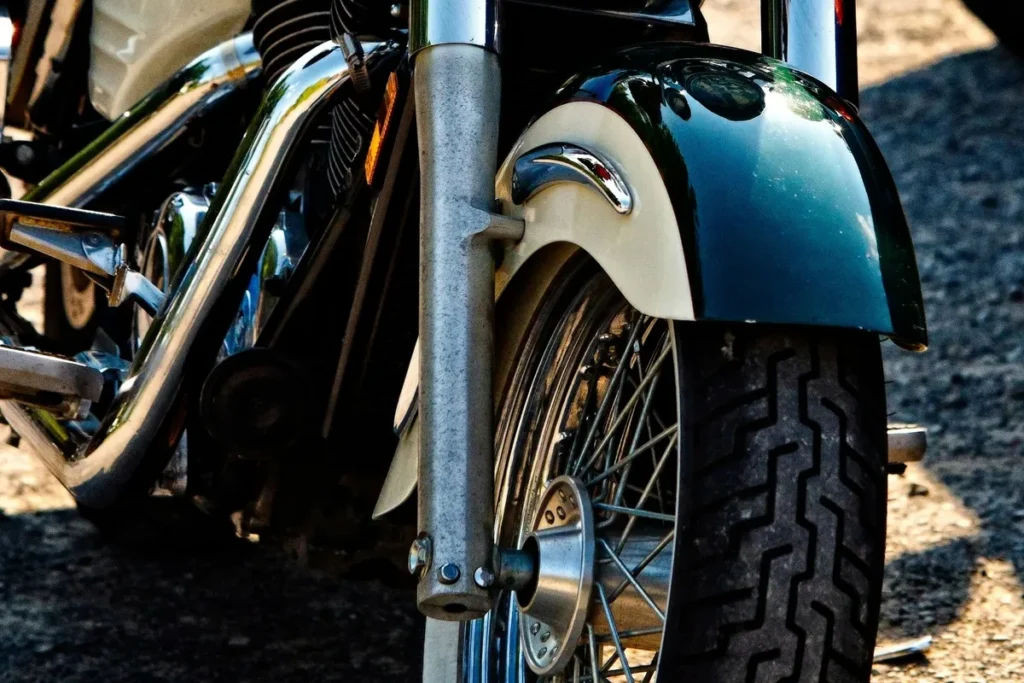 Close-up da roda dianteira de uma motocicleta estilo custom, com pneu de faixa larga, aro raiado e paralama cromado sobre estrada asfaltada.