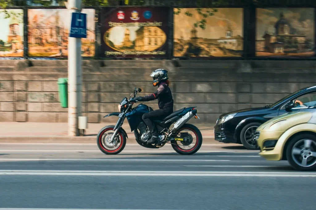 Motociclista usando capacete laranja e jaqueta preta, pilotando moto Yamaha, ao lado de carros em rua urbana.