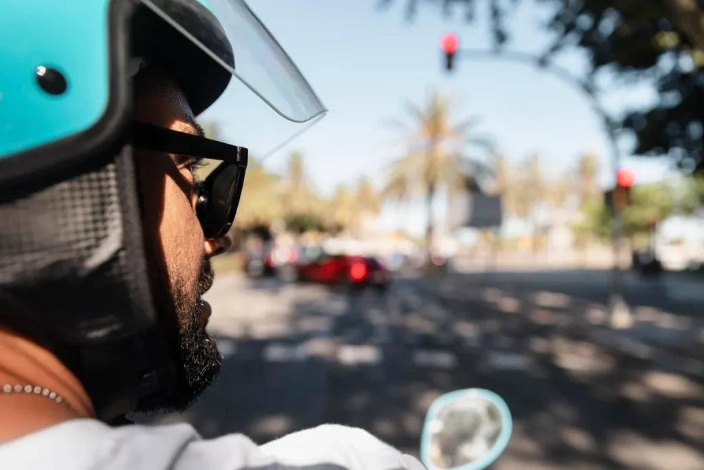 Motociclista com capacete azul e óculos escuros aguardando no sinal vermelho, em rua urbana com carros e árvores ao fundo.