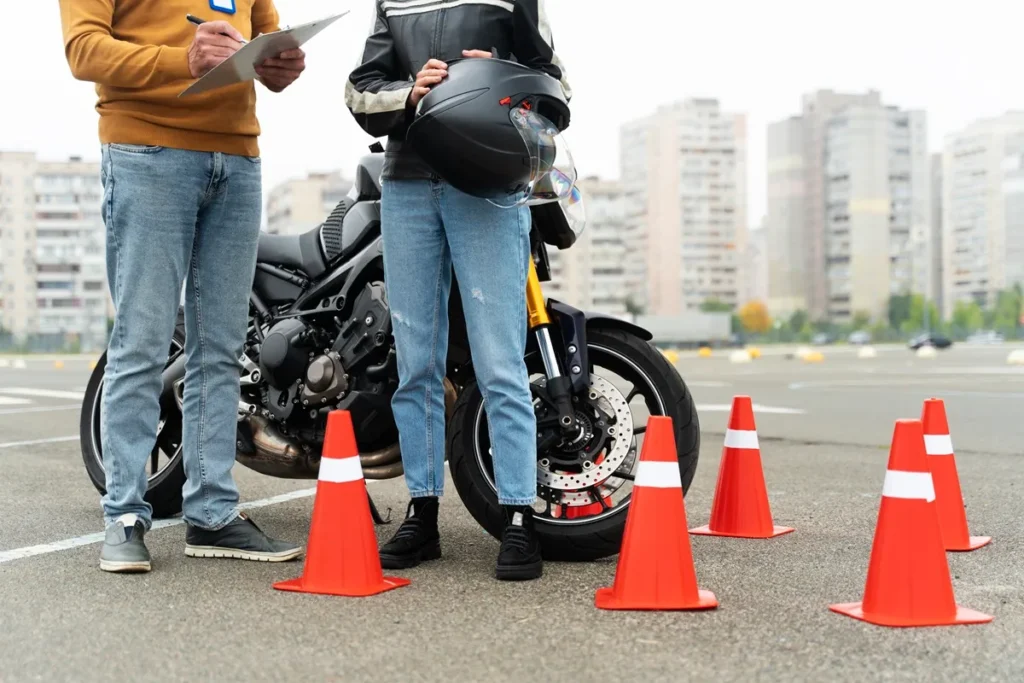 Instrutor orientando aluno em treinamento de habilitação de moto, com cones de sinalização e motocicleta no estacionamento.