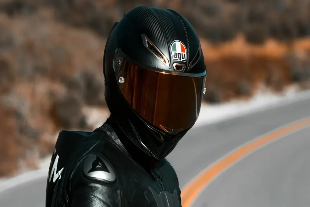 Motociclista com capacete AGV preto de viseira dourada em estrada sinuosa no deserto.