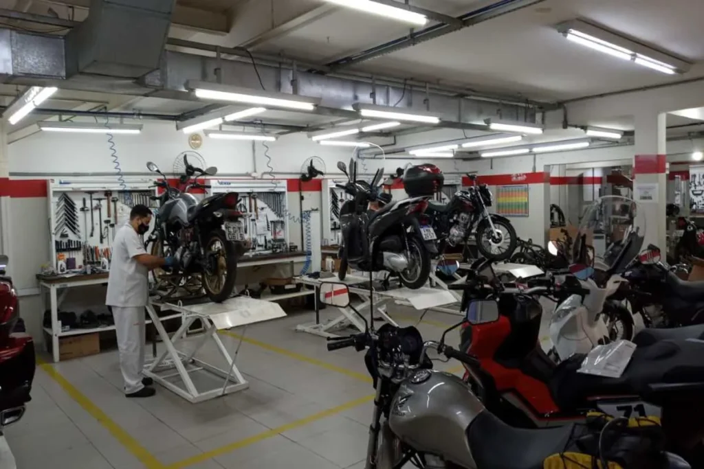 Interior de uma oficina mecânica Honda com várias motos em processo de revisão. Um mecânico de uniforme branco e máscara está trabalhando em uma moto preta elevada, enquanto outras motos, incluindo scooters, aguardam em elevadores e no chão. O ambiente é bem iluminado e organizado, com ferramentas penduradas ao fundo.