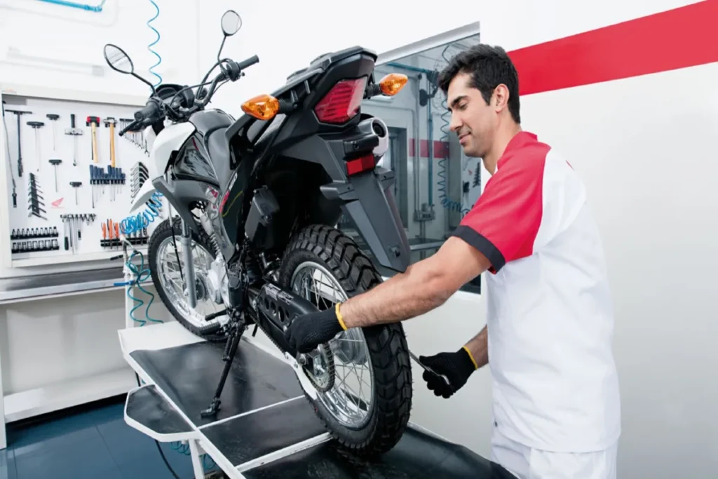 Mecânico de uniforme Honda verificando a roda traseira de uma motocicleta preta e branca em uma plataforma elevada dentro de uma oficina limpa e organizada, com painéis de ferramentas ao fundo.