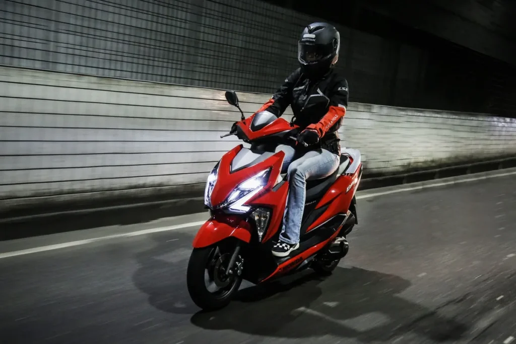 Uma pessoa pilotando uma scooter Honda Elite na cor vermelha e preta à noite, em um túnel. O motociclista usa capacete e jaqueta, e a iluminação do farol da moto destaca seu design moderno enquanto está em movimento.