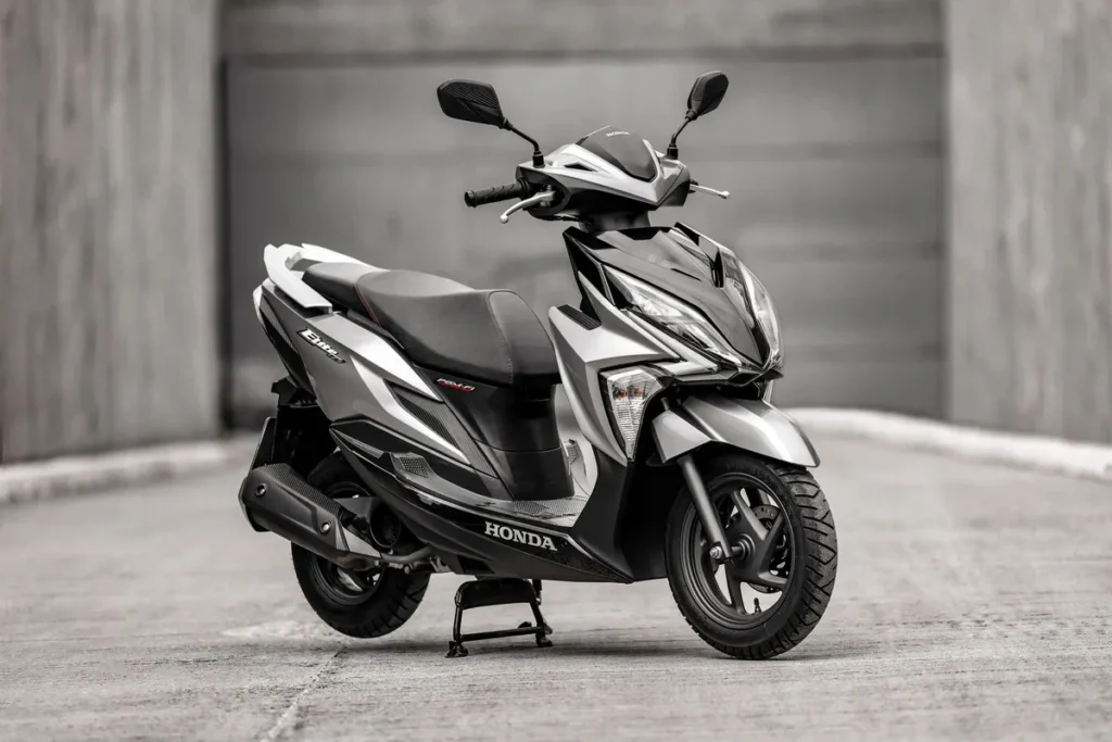 Uma scooter Honda Elite de cor prata e preta, com design moderno e angular, está estacionada em um ambiente urbano minimalista. O nome do modelo "Elite" e o logo da "Honda" são visíveis na carenagem lateral, destacando a praticidade para o dia a dia na cidade.