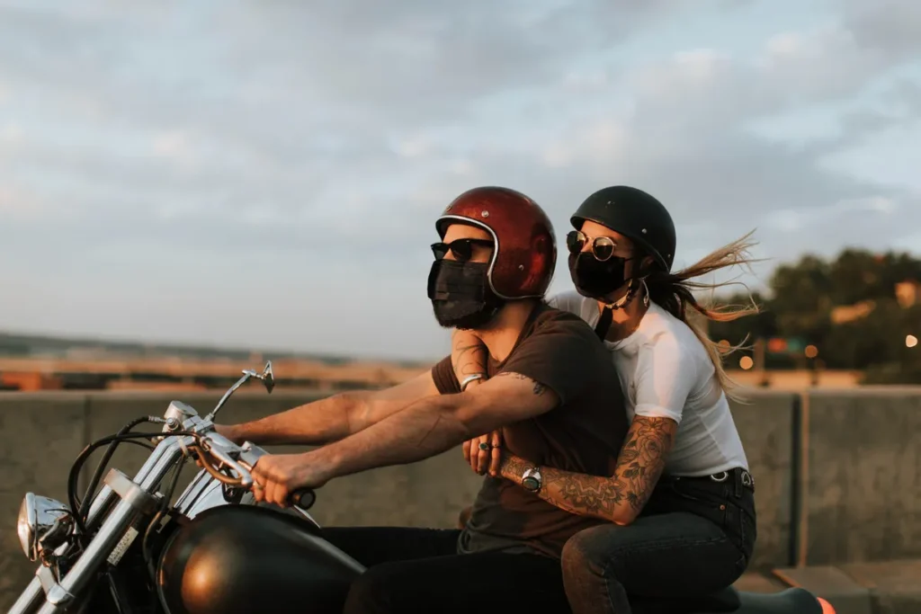 Casal de moto com a mulher na garupa, ambos usando capacetes, óculos de sol e máscaras de proteção, simbolizando uma viagem segura e moderna.