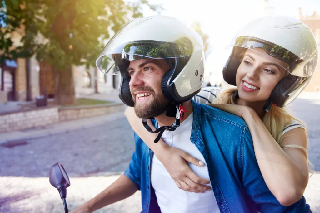 Casal sorridente em uma moto, com a mulher na garupa abraçando o homem, ambos usando capacetes brancos.