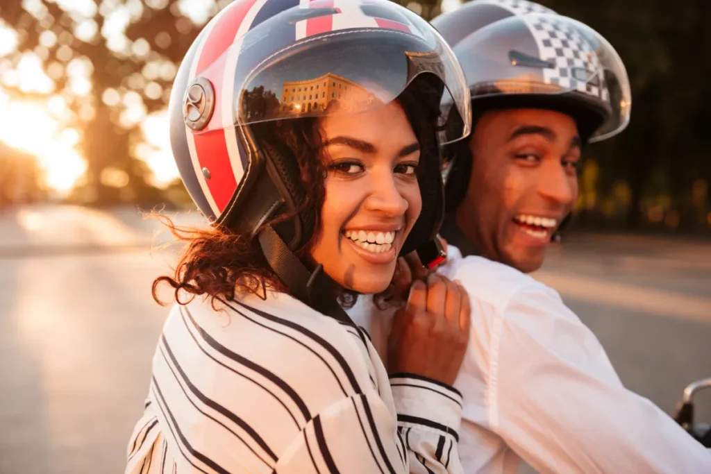 Casal sorridente em uma moto, com a mulher na garupa, ambos usando capacetes modernos com designs únicos.