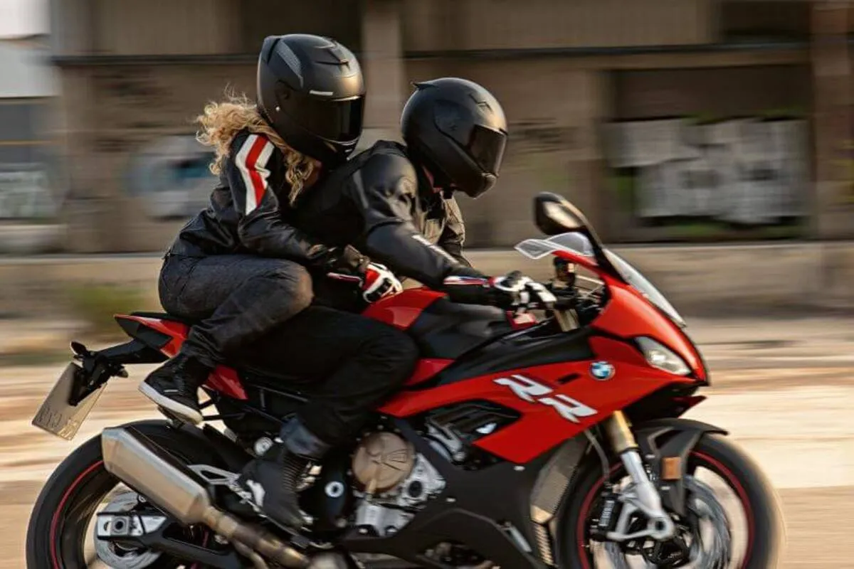 Casal com equipamentos de proteção em uma moto esportiva vermelha, com a mulher na garupa, em movimento.
