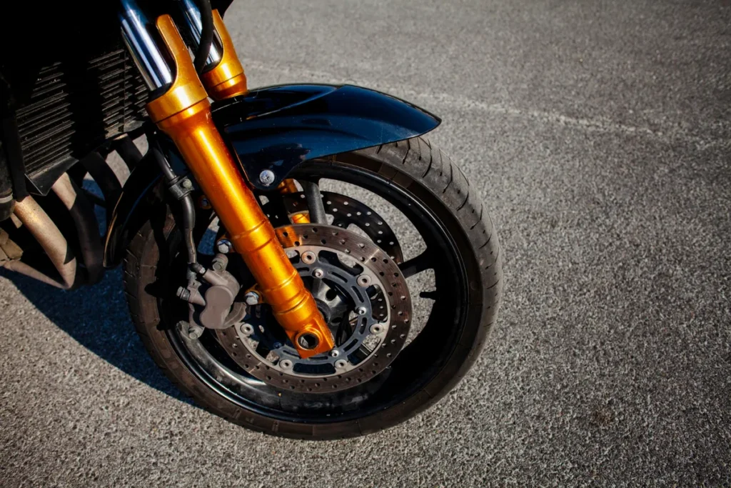 Close-up da roda dianteira de uma moto com pneu, sistema de freio a disco e suspensão dourada, sobre o asfalto.