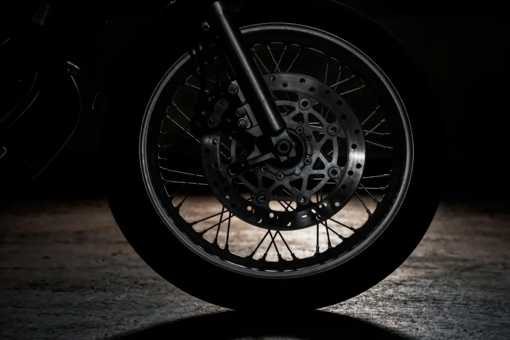 Roda de moto com raios finos e freio a disco, iluminada por uma luz forte em um fundo preto.
