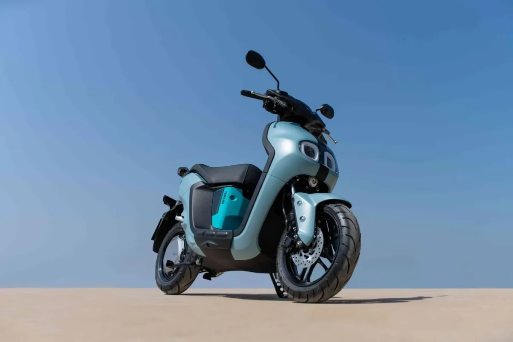 Scooter elétrica moderna, na cor azul-claro/verde-água, estacionada em piso de concreto sob céu azul. Veículo de mobilidade urbana e design minimalista.