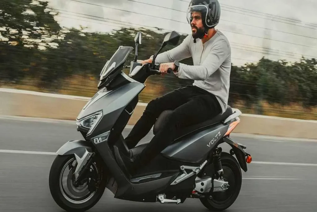 Homem de capacete e camisa clara a conduzir uma scooter elétrica cinza-escura numa estrada. Cena de mobilidade urbana e transporte moderno.