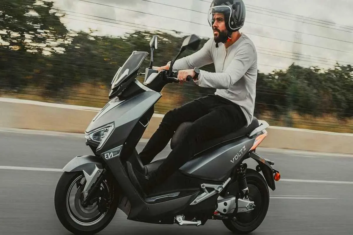 Homem de capacete e camisa clara a conduzir uma scooter elétrica cinza-escura numa estrada. Cena de mobilidade urbana e transporte moderno.