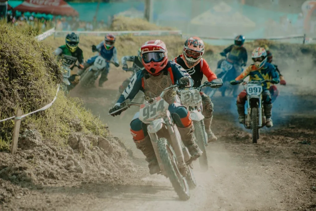 Pilotos competindo em rally motos em uma pista de terra com curvas e poeira