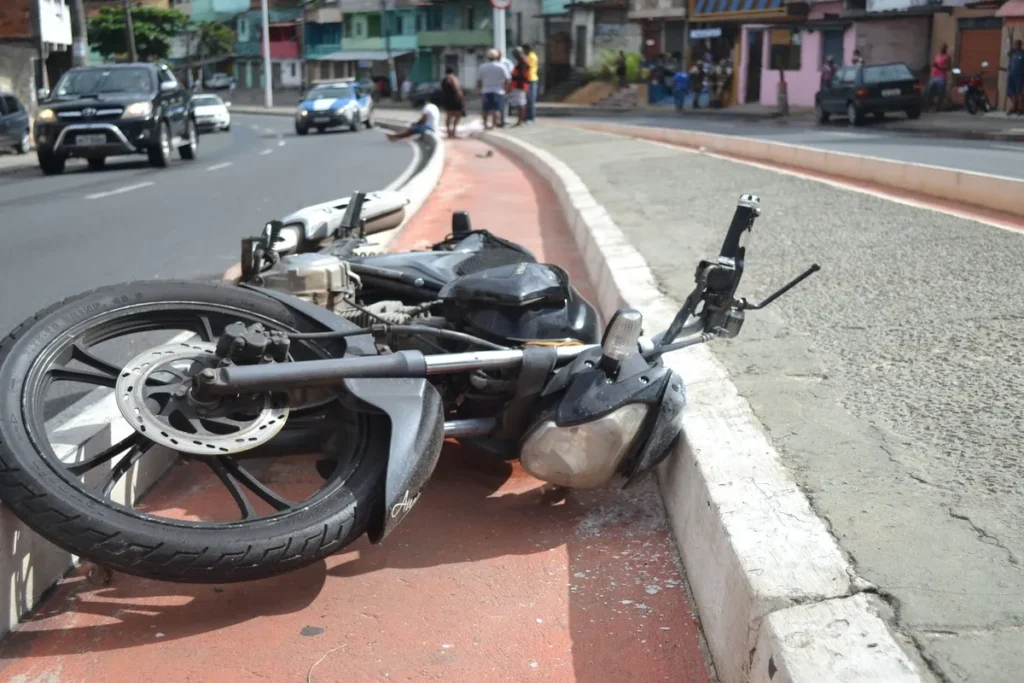 Uma motocicleta escura, provavelmente de baixa cilindrada, está caída de lado em uma área de ciclovia pintada de vermelho, encostada no meio-fio de uma rua urbana. A moto está danificada, com a carenagem e o farol frontais quebrados. Em segundo plano, há tráfego de carros, um veículo da polícia (em azul e branco) e pessoas aglomeradas na calçada, indicando um recente acidente de trânsito.