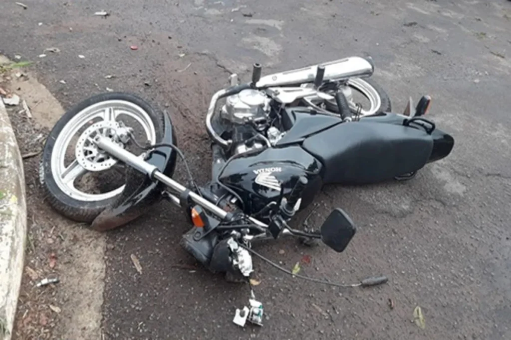 Vista em close-up de uma motocicleta preta (modelo Honda, provavelmente) caída e tombada na lateral em uma rua de asfalto. A moto está danificada na parte frontal, com peças e fiação soltas perto do guidão. O pneu dianteiro está no chão ao lado do meio-fio e terra. A cena é de um acidente de trânsito ou queda de moto.