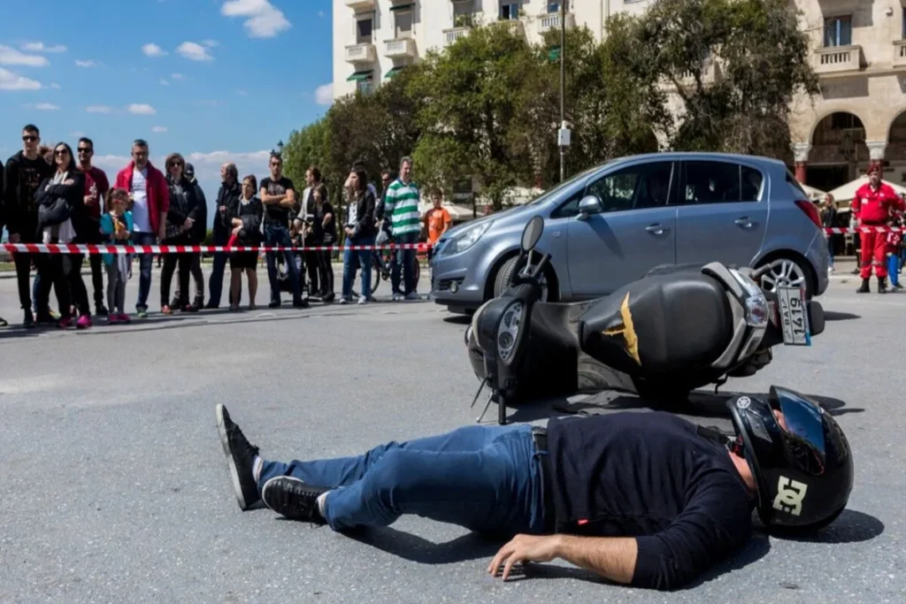Um motociclista, vestindo calças jeans e uma camisa escura, está deitado de lado no asfalto com o capacete ainda na cabeça, após uma queda de moto. Uma scooter ou moto preta está tombada ao lado dele, e um carro cinza está parado por perto. Um cordão de isolamento de segurança e uma multidão de pessoas observando a cena completam o plano de fundo em um dia ensolarado.