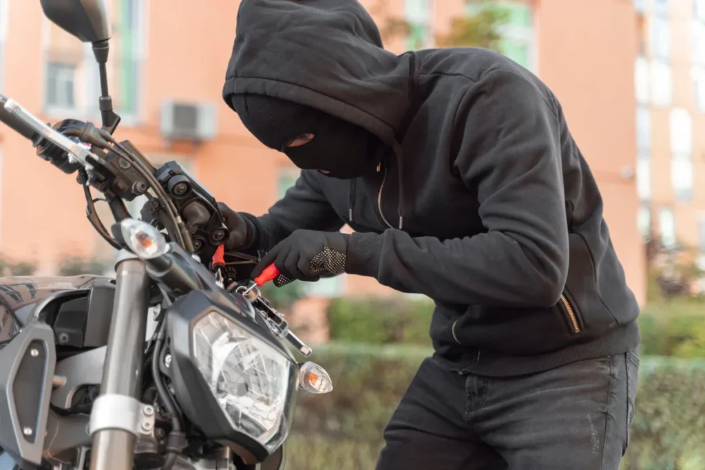 Um indivíduo vestido com moletom preto de capuz e balaclava preta (máscara), cobrindo completamente o rosto, está inclinado sobre a área do guidão de uma motocicleta escura. Ele usa luvas e está segurando uma ferramenta vermelha (possivelmente uma chave de fenda ou alicate) enquanto mexe nos fios e na ignição da moto, simulando uma tentativa de furto. O fundo mostra um prédio de cor pêssego e folhagem verde.