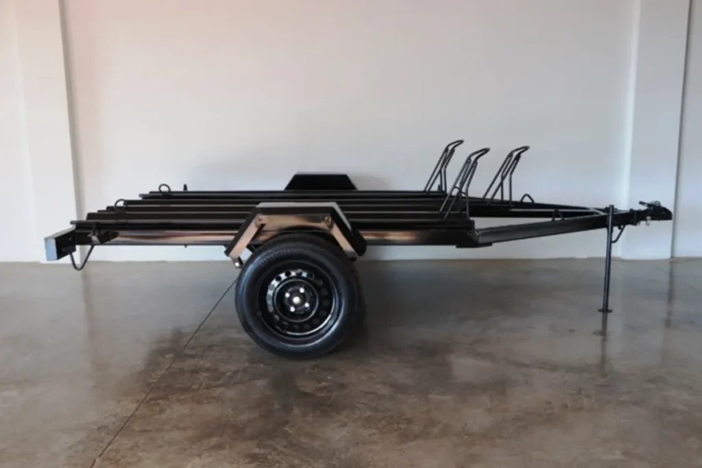 Um reboque moto de cor preta e eixo único, vazio, visto de lado em um estúdio. O reboque é projetado para transportar até três motocicletas, com os suportes de roda verticais visíveis.
