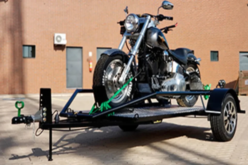 Uma motocicleta custom ou chopper prateada e preta está firmemente presa em um reboque moto preto, com cintas verdes e prateadas, em frente a um prédio de tijolos.