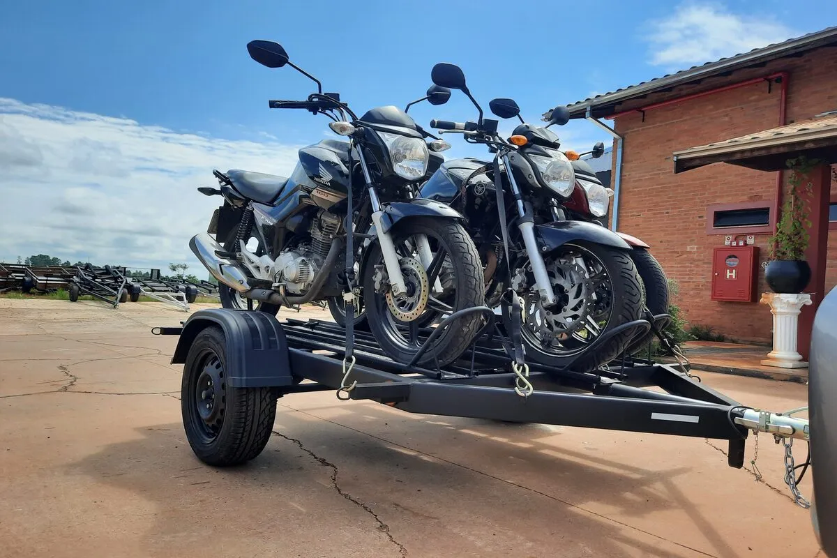 Um reboque moto preto, engatado a um veículo, transporta duas motocicletas de baixa a média cilindrada em um dia ensolarado. As motos pretas estão seguras com cintas de amarração visíveis na plataforma.