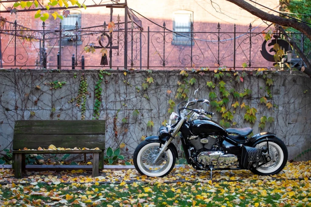 Moto bobber preta estacionada ao lado de um banco de madeira em um jardim com folhas secas